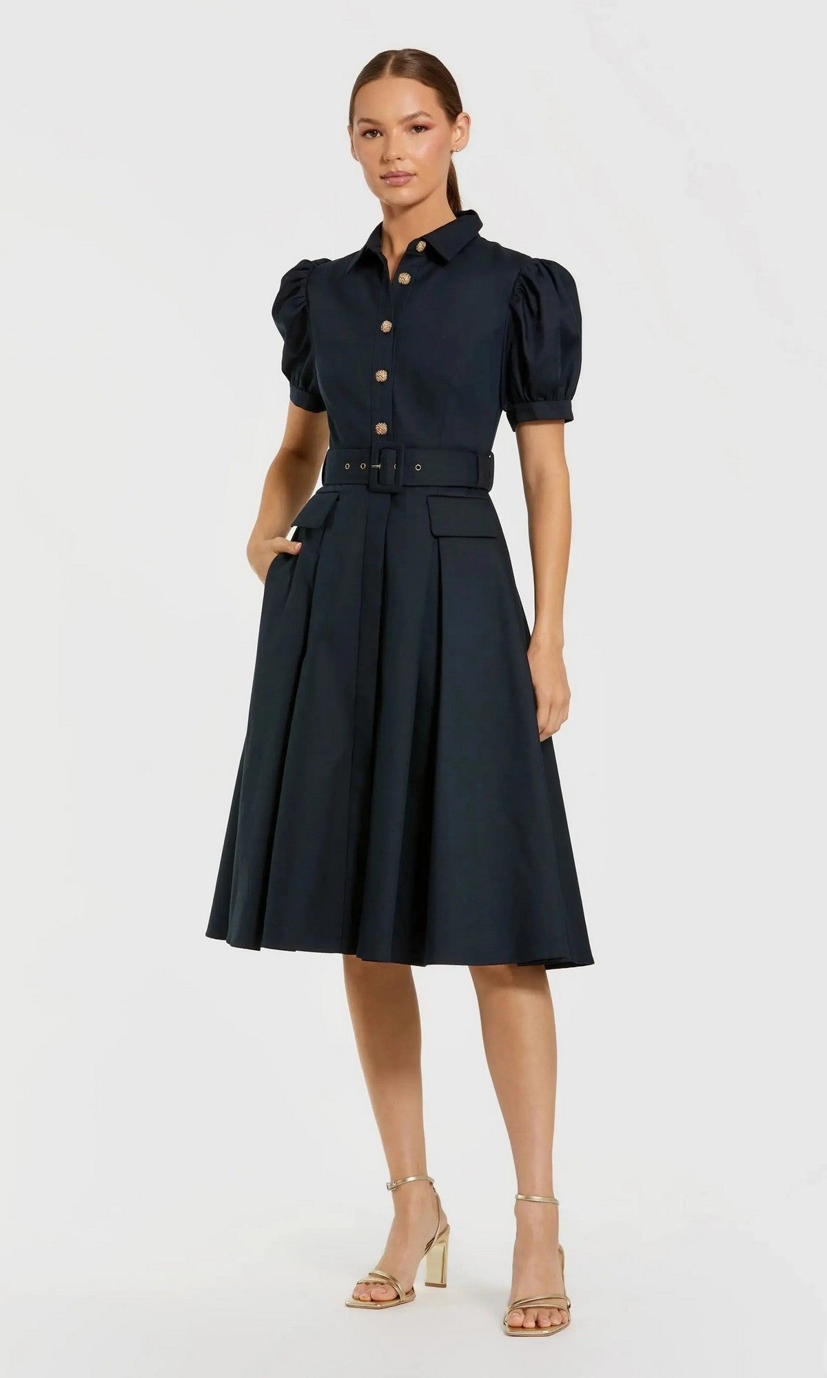 A woman stands wearing the Mac Duggal 69097 Knee Length Belted Shirt Dress in Midnight, featuring a collared neckline, button-down front, puff sleeves, and an elegant a-line skirt. She completes the look with gold strappy heels against a plain light background.
